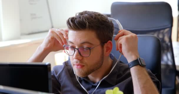 ヘッドセットを着用し 近代的なオフィスのデスクで働く青年の白人実業家のフロント ビュー 彼は音楽を聴く — ストック動画
