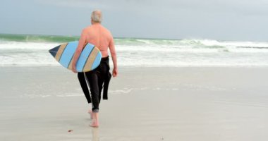 Dikiz Plajı'nda sörf tahtası ile yürüyen yaşlı beyaz adam. Dalgalar deniz kıyısı içinde belgili tanımlık geçmiş 4k üzerinde
