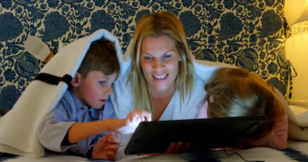 彼女の息子とデジタル タブレットを使用して 自宅の寝室のベッドの上の娘と若い白人の母親のフロント ビュー の毛布の下 します — ストック動画