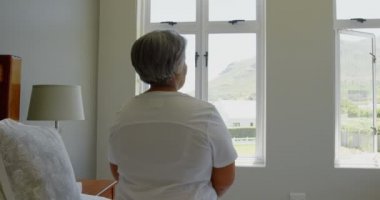 Siyah kadın yatakta rahat ev yatak odasında oturmuş kıdemli görünümünü arka. 4 k pencereden dışarı bakıyor