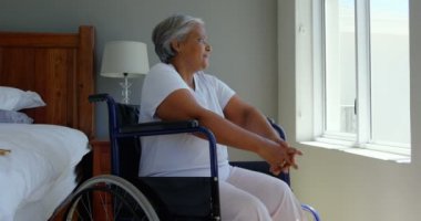 Evde yatak odasında pencere yakınındaki tekerlekli sandalye üzerinde oturan handikap siyah kadın kıdemli yan görünüm. 4 k pencereden dışarı bakıyor