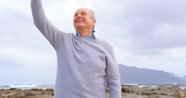 ビーチでの携帯電話で Selfie を取って古い白人の年配の男性のフロント ビュー 美しい空と山々 を背景 — ストック動画