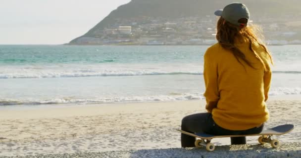 年轻的高加索妇女坐在滑板上 看着海滩上的大海的后景 妇女在海滩在阳光4K — 图库视频影像