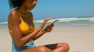 Güzel genç Afro-Amerikan kadın plajda güneş altında dijital tablet kullanarak yan görünüm. 4 k sahilde rahatlatıcı