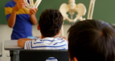 Front view of mixed-race student explaining skeleton model in classroom at school. Classmates listening to him 4k