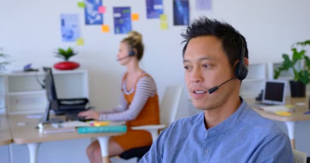 Vista Lateral Joven Ejecutivo Asiático Hablando Con Auriculares Escritorio Caucásico — Vídeo de stock