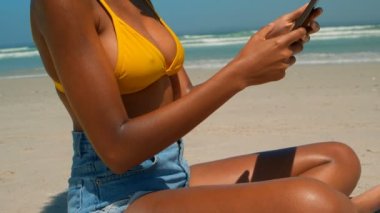 Güzel genç Afro-Amerikan kadın plajda güneş altında dijital tablet kullanarak yan görünüm. Orta bölüm 4k sahilde rahatlatıcı kadının