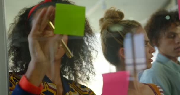 つのクラスメート Inbackground と近代的なオフィスに付箋の若いアフリカ系アメリカ人の女性エグゼクティブ作業のフロント ビュー — ストック動画