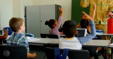 Okul çocuklarının çeşitli grubun arka görünümü, sınıf 4k 'da yangın güvenliği konusunda Kafkas erkek itfaiyecinin öğretilmesi sırasında el yükselterek