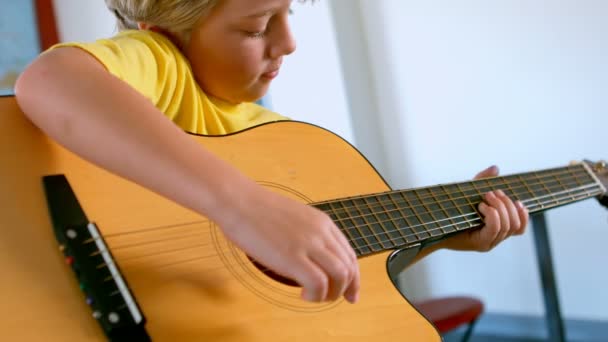 かわいいブロンドの白人少年の机に座って 教室でギターを弾くの側面図です 彼は学習ギター — ストック動画