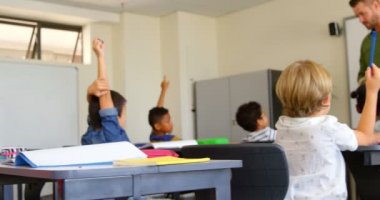 Okulda bir sınıfta masada otururken çok etnik okul çocuklarının el yükselterek yan görünümü. Erkek öğretmen ellerini 4k yükselterek öğrencilere işaret