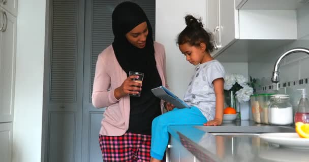 在家里4K 的厨房里 一名年轻的亚洲母亲拿着数字平板电脑向女儿拿着数字平板电脑 一边喝着一杯水的年轻亚洲母亲的面前 — 图库视频影像