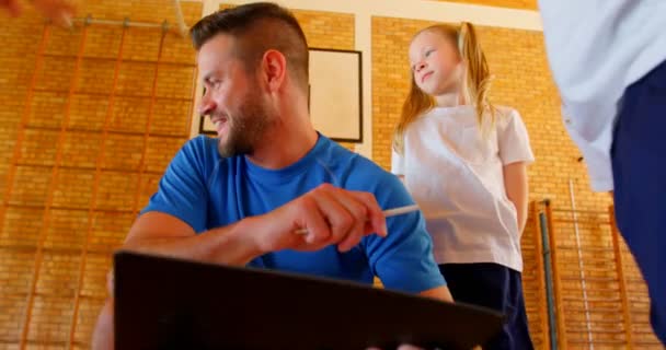 Vue de face du jeune professeur de sport caucasien et des écoliers discutant sur le presse-papiers dans le terrain de basket-ball. Ils écoutent le professeur de sport 4k 