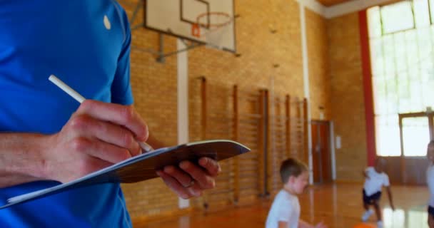 白人男性バスケットボールコーチのミッドセクション学校のバスケットボールコートでメモ帳に書いています 背景4K で遊んで学童達 — ストック動画