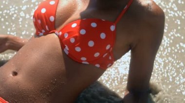 Kırmızı bikini beyaz lekeli dalgalar, sahilinde yalan ile olan Afrika kökenli Amerikalı kadın önden görünümü. Orta bölüm 4k ışığında sahilde rahatlatıcı kadının