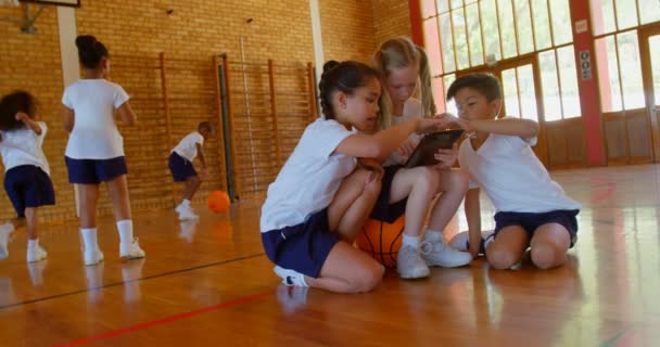 在学校的篮球场上 使用数字平板电脑的一群不同的小学生的正面观点 其他孩子在后台玩4K — 图库视频影像