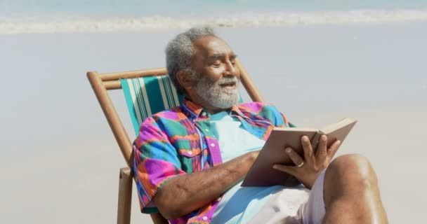 活跃在海滩的躺椅上看书的活跃非洲裔美国老人的正面观点 他微笑着4K — 图库视频影像