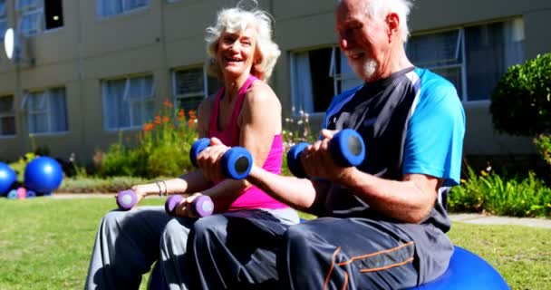 老人ホームの庭でダンベルで運動をする白人の高齢者のカップルの側面図 彼らはお互いに相互作用している4K — ストック動画