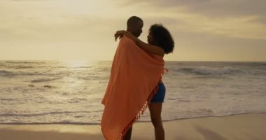 Sahilde battaniyeye sarılmış Afro-Amerikan çiftin yan görünümü. Birbirlerini kucaklıyorlar 4k