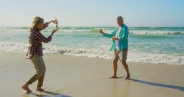 Sahilde cep telefonu ile üst düzey adamın fotoğrafları tıklayarak aktif üst düzey Kafkas kadın Yan görünümü. Onlar eğlenceli 4k yaşıyorsanız