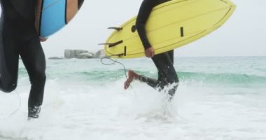 Sahilde sörf tahtasıyla koşan iki erkek sörfçünün alçak kesimi. Arka planda deniz dalgaları 4k
