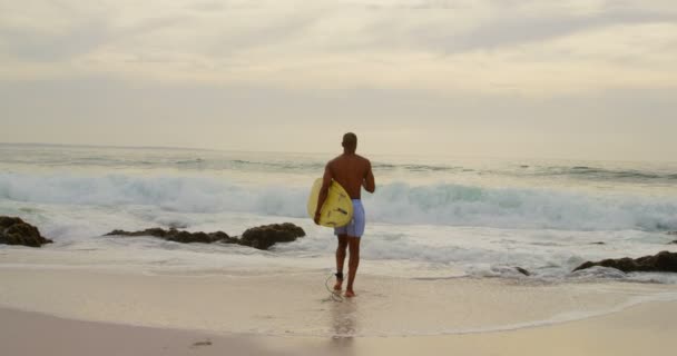 非洲裔美国男性冲浪者在海滩上用冲浪板奔跑的后视图 平静的海在背景 — 图库视频影像