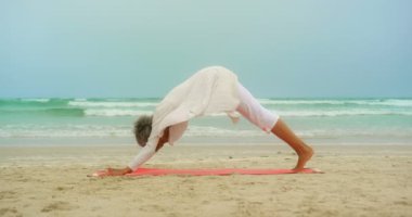 Plajda egzersiz mat yoga yapan aktif yaşlı Afro-Amerikan kadının yan görünümü. O adho mukha svanasana 4k yapıyor