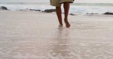 Güneşli bir günde sahilde yalınayak yürüyen bir adam düşük bölümü. Arka planda deniz dalgaları 4k