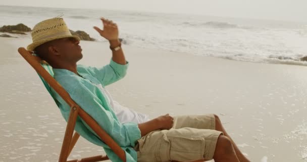 在海滩上的日光浴躺椅上放松的非洲裔美国人的侧视图 他看着远方4K — 图库视频影像