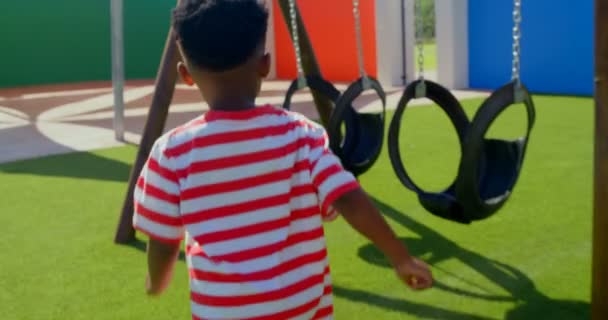 学校の遊び場でスイングに向かって走っているアフリカ系アメリカ人の男子生徒の背面図 彼は晴れた日4Kで遊んでいます — ストック動画
