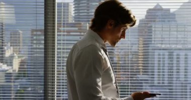 Genç Kafkas erkek yöneticinin modern ofiste pencerenin yanında cep telefonuyla konuşmasının yan görünümü. Pencereden 4k'ye bakıyor.