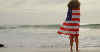 Sahilde Amerikan bayrağına sarılı Afro-Amerikan kadının arka görünümü. O ayakta ve deniz 4k bakıyor