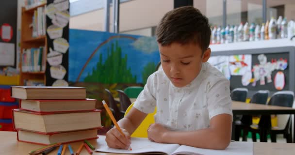 学校の教室で机の上で勉強しながら手を挙げるアジアの男子学生の正面図 彼はノートに書いている — ストック動画