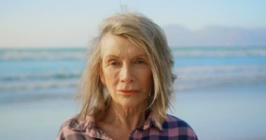 Sahilde duran aktif kıdemli Beyaz kadının ön görünümü. O gülümsüyor ve kamera 4k bakıyor