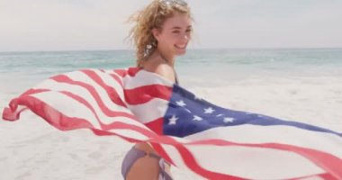 Sahilde Amerikan bayrağı sallayarak beyaz bir kadının ön görünümü. O gülümsüyor ve kamera 4k bakıyor