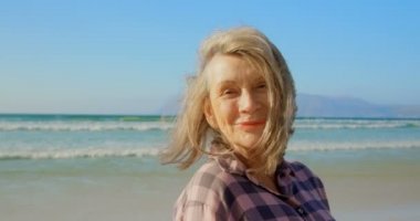 Sahilde duran aktif kıdemli Beyaz kadının ön görünümü. O gülümsüyor ve kamera 4k bakıyor