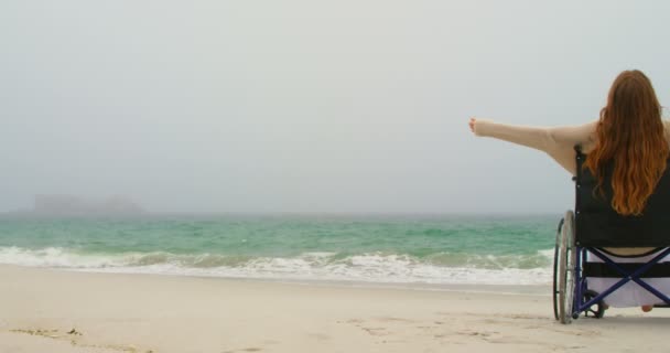 年轻的白种妇女坐在沙滩上的轮椅上 双臂伸出的侧视图 她感觉幸福4K — 图库视频影像