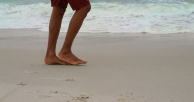 Güneşli bir günde sahilde yalınayak yürüyen bir adam düşük bölümü. Arka planda deniz dalgaları 4k