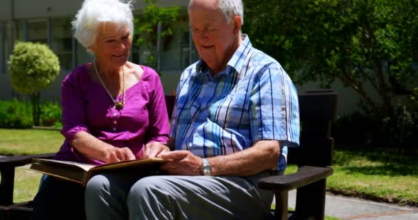 活跃高加索老年夫妇在疗养院的花园里看相册的正面视图 他们坐在花园里 — 图库视频影像