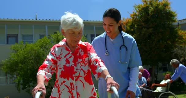 老人ホームの庭で歩行者と一緒に歩くのを助ける白人女性医師の正面図 バックグラウンドの高齢者 — ストック動画