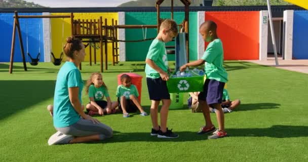 学校の遊び場でボトルのリサイクルについて学校の生徒を教える白人女性教師の側面図 遊び場に座っている小学生 — ストック動画