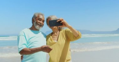 Sahilde cep telefonu ile selfie çeken aktif üst düzey Afro-Amerikan çift ön görünümü. Onlar gülümsüyor ve 4k konuşuyor
