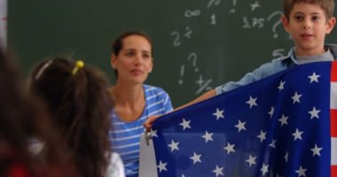 Beyaz öğrencinin sınıfta Amerikan bayrağını açıklamasının ön görünümü. Arka planda oturan kadın öğretmen 
