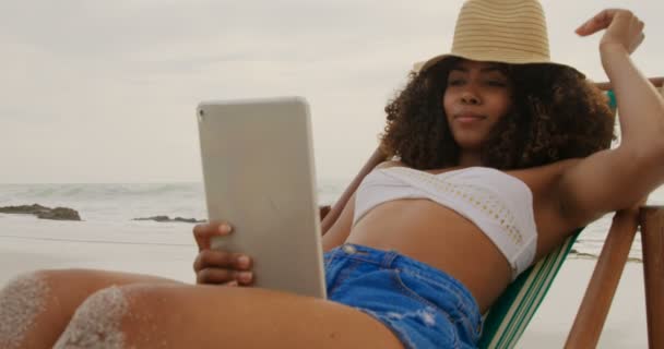 ビーチでデジタルタブレットを使用してアフリカ系アメリカ人女性の正面図 彼女はサンラウンジャー4Kでリラックスしています — ストック動画