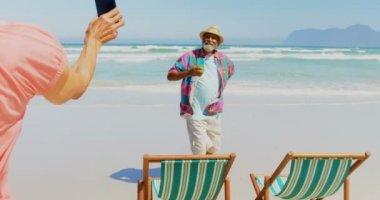 Sahilde cep telefonu ile adam fotoğrafı çeken aktif üst düzey Afro-Amerikan kadın Arka görünüm. Kokteyl bardağı 4k ile poz adam