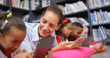 Okul kütüphanesinde dijital tablet üzerinde okuyan Kafkas kadın öğretmen ve okul çocuklarının ön görünümü. Kız öğrenci ile etkileşim kadın öğretmen 
