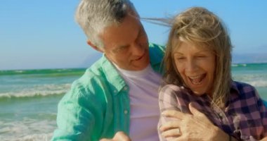 Sahilde cep telefonu ile selfie çeken aktif üst düzey Kafkas çiftin ön görünümü. Birbirlerini kucaklıyorlar 4k