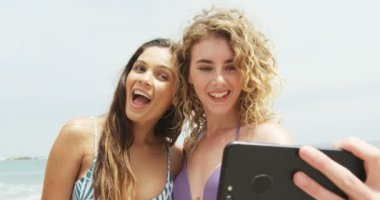 Sahilde cep telefonu ile selfie çeken iki Karışık ırklı kadın arkadaşın ön görünümü. Onlar gülümsüyor ve eğleniyor4k