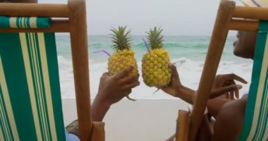 Sahilde ananas suyu kızartma Afrikalı Amerikalı çift in arka görünümü. Onlar bir şezlong 4k üzerinde rahatlatıcı 