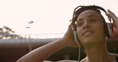 Şehirde kulaklıkla müzik dinleyen genç Afro-Amerikan kadının ön görünümü. O gülümsüyor ve uzağa 4k bakıyor